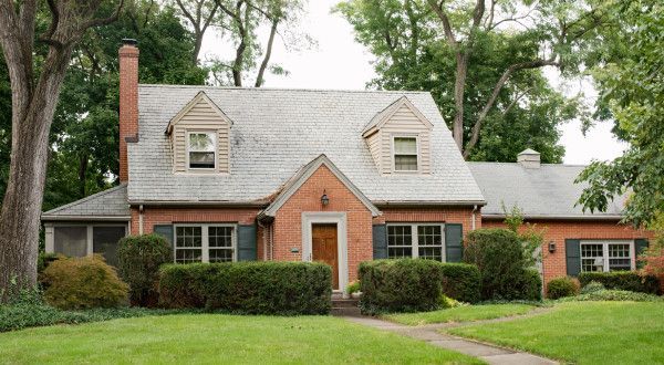 Brick house with gray roof, central red door, and manicured lawn amid tall trees