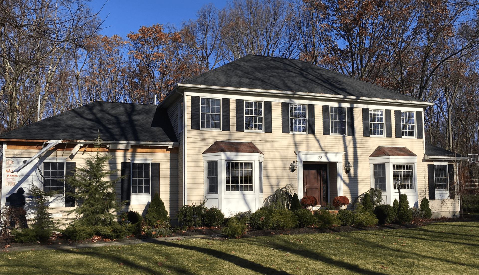 Two-story beige suburban house with dark roof, white trim, and bare trees in front on a sunny day