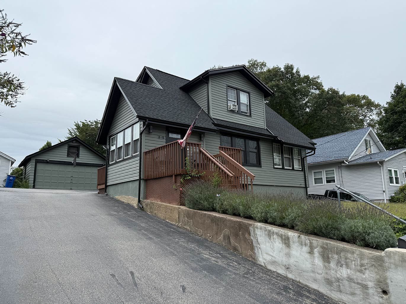 Large gray house on a sloped driveway with a wooden staircase and small outbuildings nearby