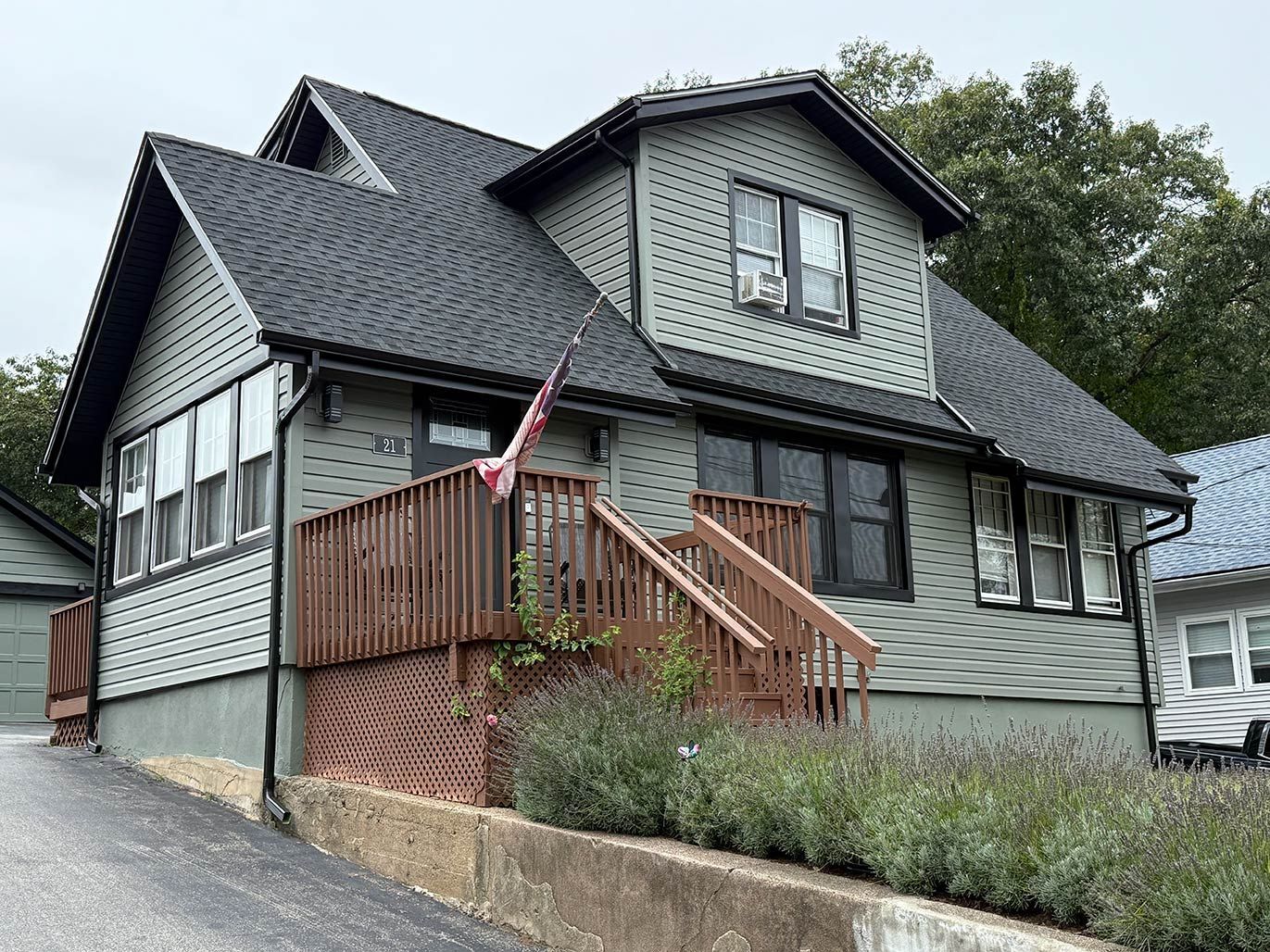 Gray house with black roof, wooden porch, and an American flag along a driveway.