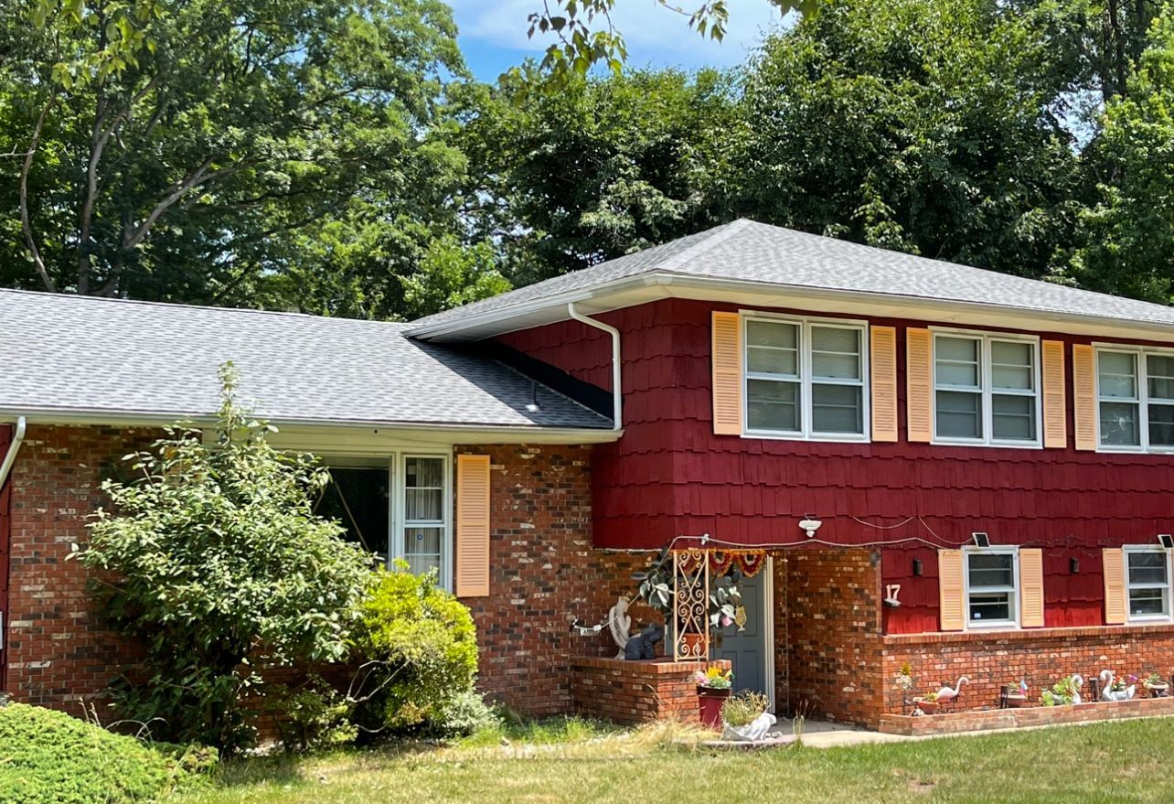 Two-story red brick and burgundy house with gray roof, white-trim windows, and trees behind it