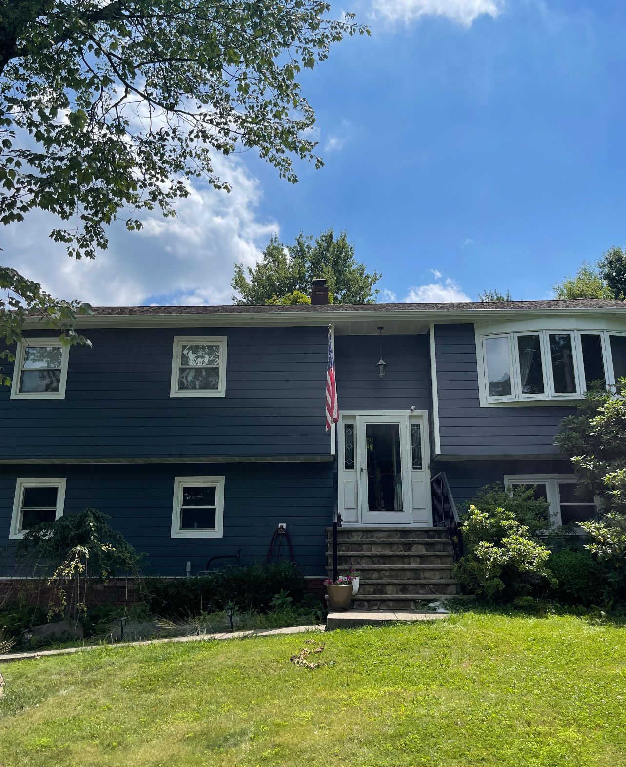Dark gray two-story house with white trim, front steps, and an American flag under a blue sky.