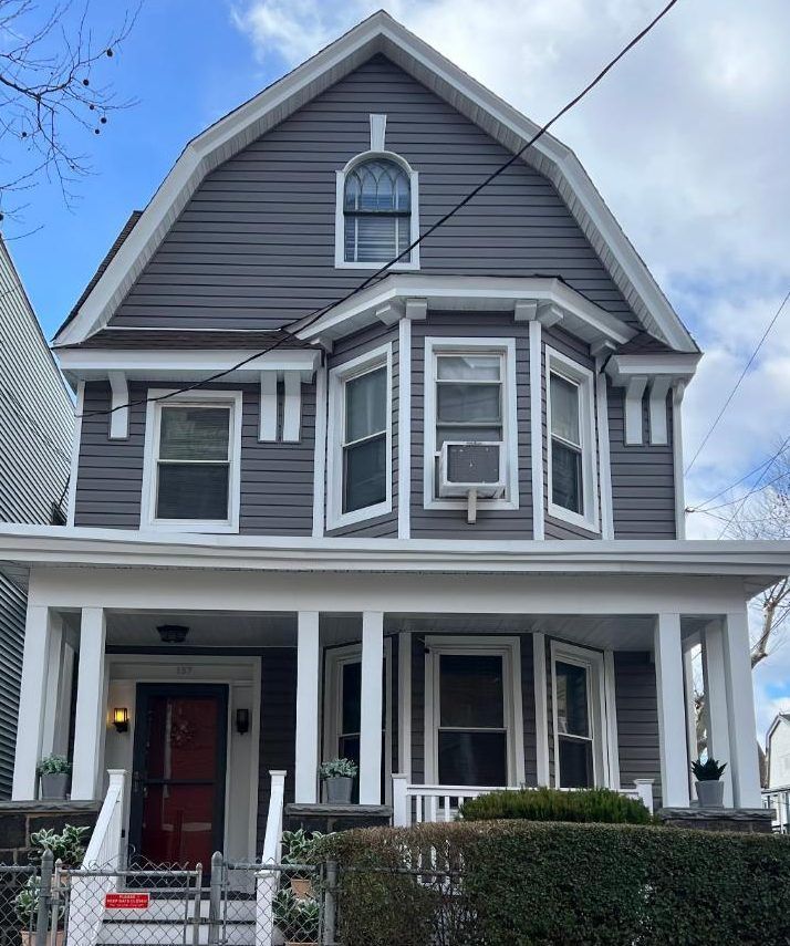 Gray Victorian house with white trim, front porch, and red door under a cloudy sky