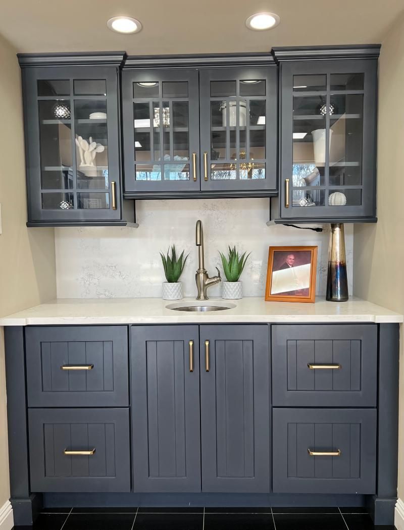 Gray built-in cabinet with glass-front upper doors, white countertop, and small plants beneath recessed lights
