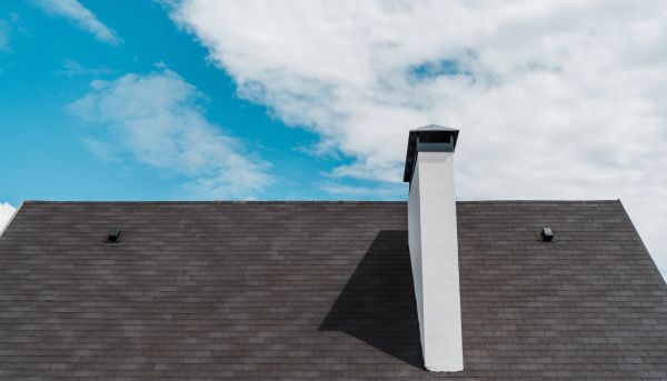 Roof with dark shingles and a white chimney against a blue, cloudy sky