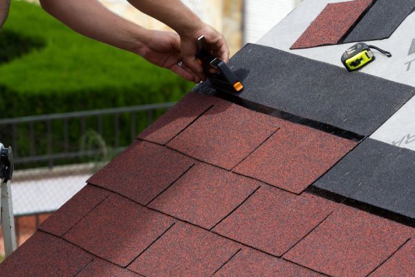 Hands using a utility knife to cut black roofing felt on a red shingled roof