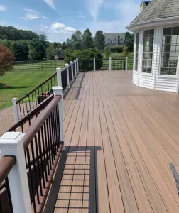 Wooden deck with black railing beside a white house, overlooking a green lawn and trees under a blue sky