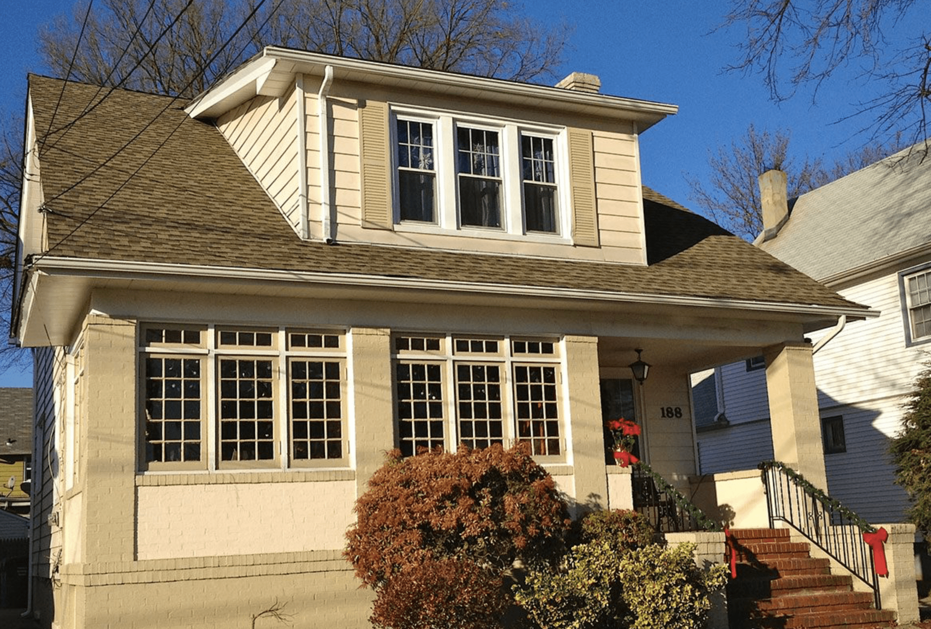 Small beige house with a front porch, dormer windows, and shrubs under a clear blue sky