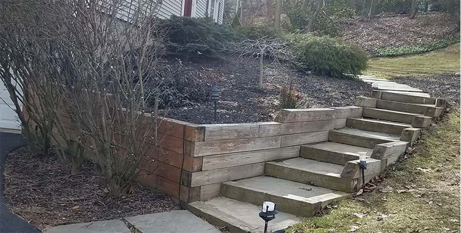 Landscaped retaining wall with stone steps leading up a sloped yard beside a sidewalk.