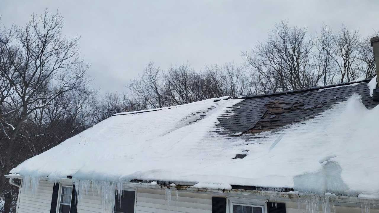 Snow-covered house roof with a large dark tarp patch and bare trees in the background