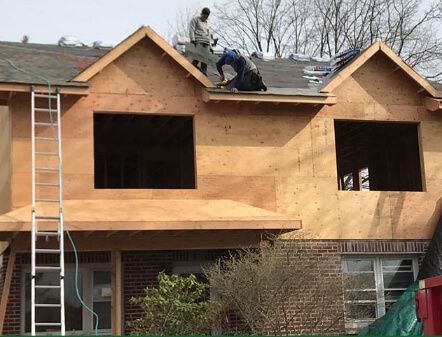 Workers reroofing a house with exposed plywood and dormer windows.
