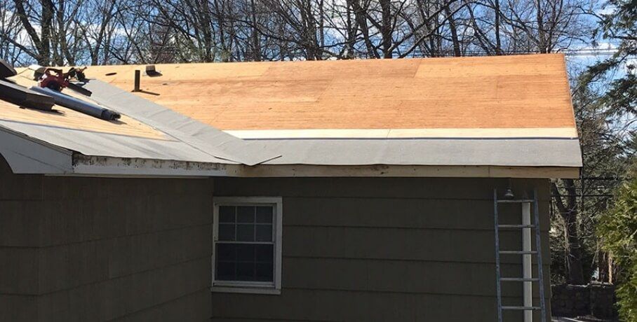 House roof under repair with exposed plywood and white underlayment beside gray siding and windows