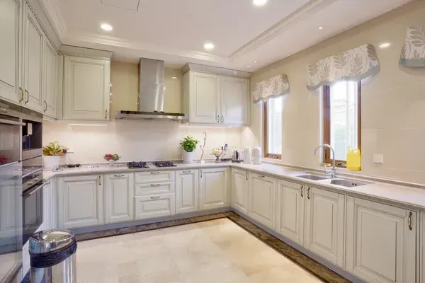 Bright cream U-shaped kitchen with white cabinets, marble counters, stainless range hood, and window valances.