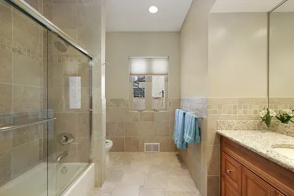 Bright beige bathroom with glass shower, tiled floor, window, and wooden vanity with sink