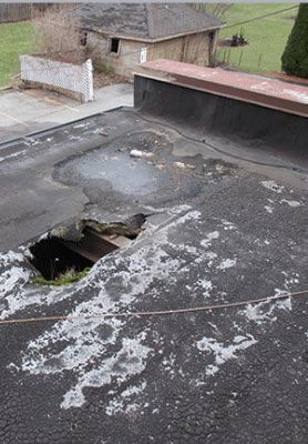 Damaged flat roof with a large hole, peeling membrane, and nearby chimney in a rural setting