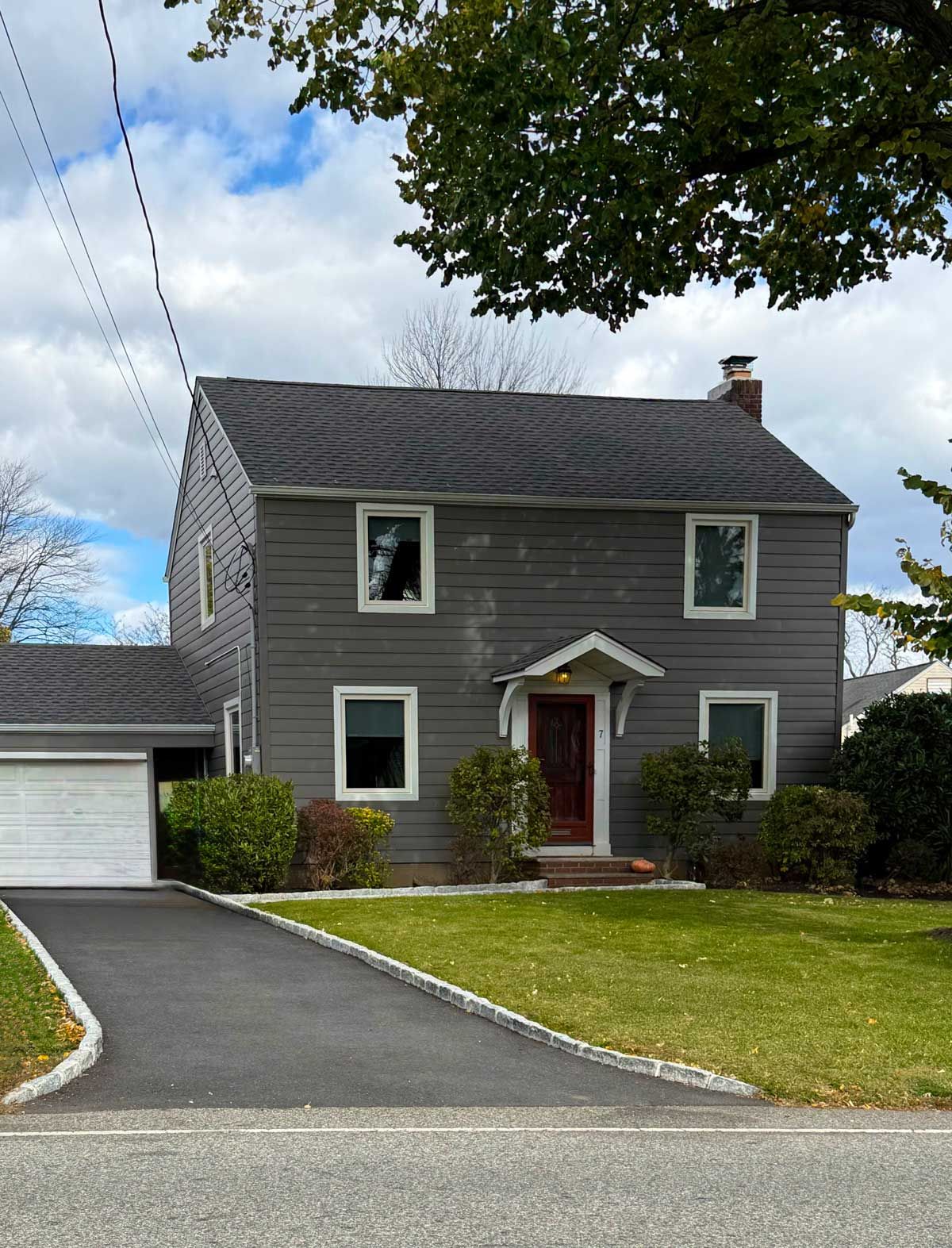 Gray two-story house with a red front door, driveway, lawn, and tree branches overhead