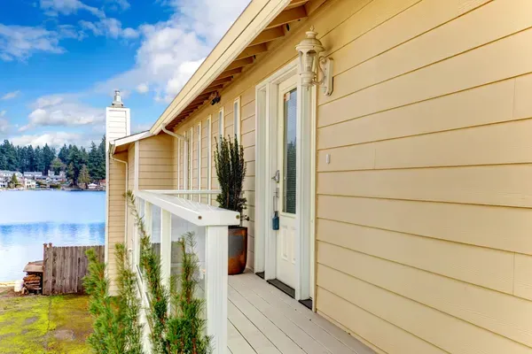 Yellow lakeside house porch with white door and railing beside calm water under a blue sky