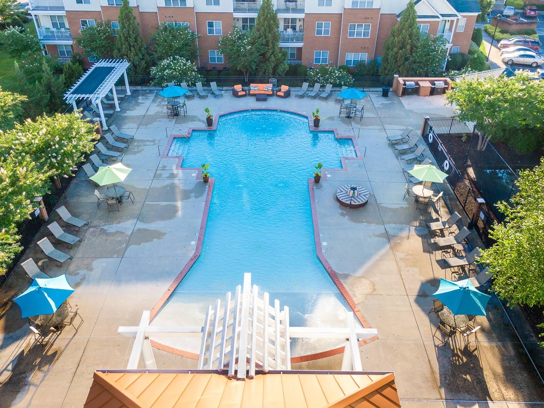 An aerial view of a swimming pool shaped like a keyhole, surrounded by lounge chairs, umbrellas, and a patio area.