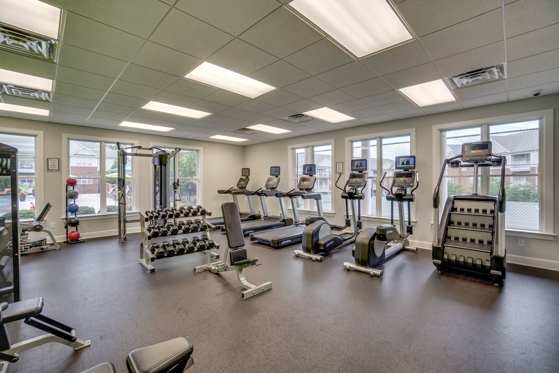 A clean, modern apartment gym featuring a row of treadmills, elliptical machines, dumbbell racks, and benches.