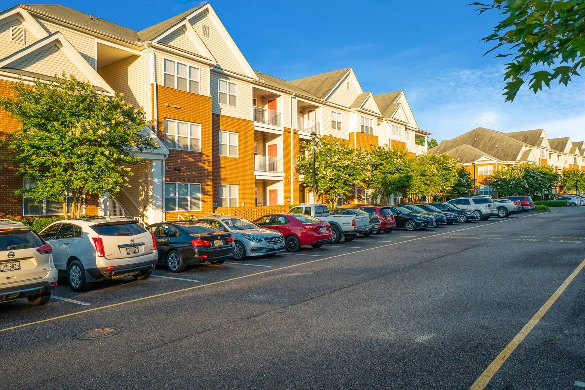 A multistory apartment complex with tan and brick siding, fronted by a parking lot filled with various cars on a sunny day.