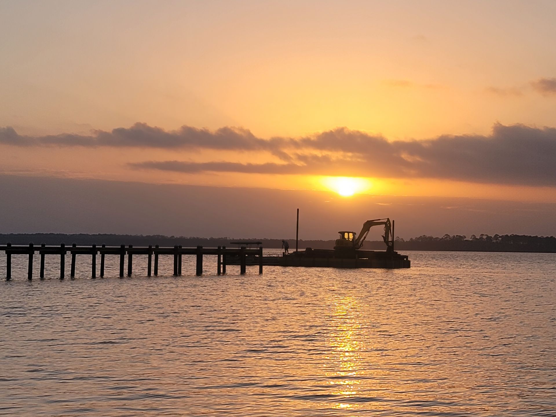 Sunset over water with a pier and construction equipment; orange and yellow sky.