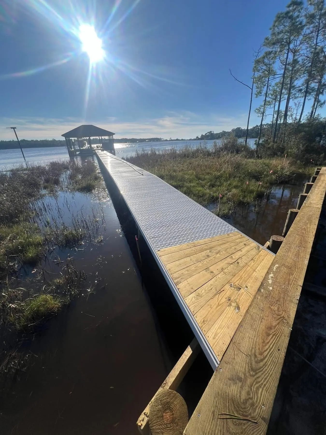 A long wooden pier extends over dark water, leading to a gazebo under a sunny sky.