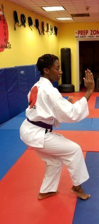 a woman in a karate uniform is squatting on a mat in a gym .