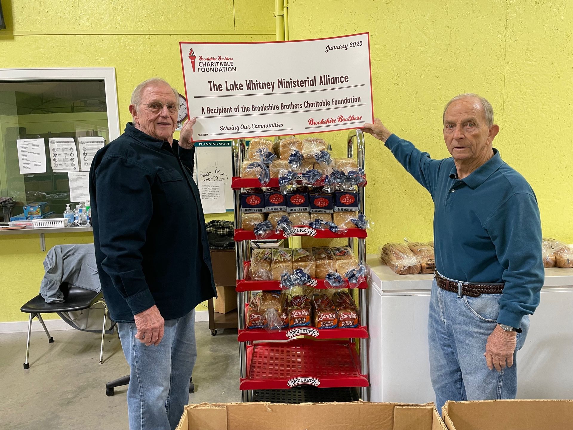 Two men are standing next to each other holding a check in front of a display of bread.