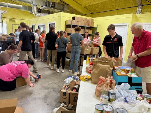 Volunteers pack food boxes in a brightly lit warehouse. People sort, bag, and organize donations.