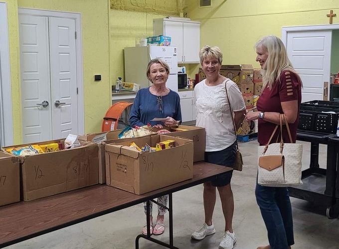 Three women are standing around a table filled with boxes of food.