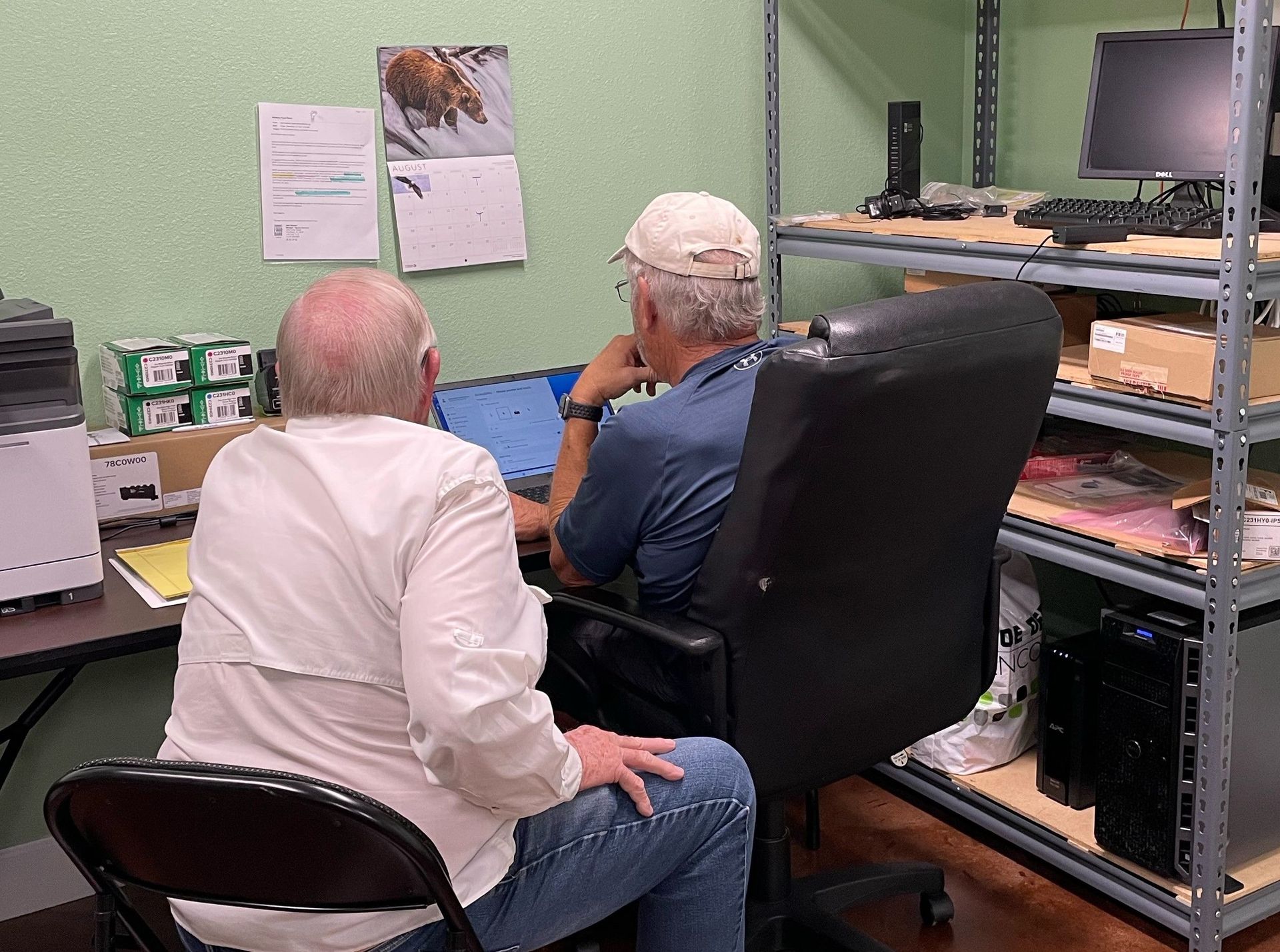 Two men are sitting in chairs in front of a computer.