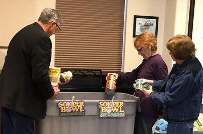 A group of people are standing around a soup bowl in a room.