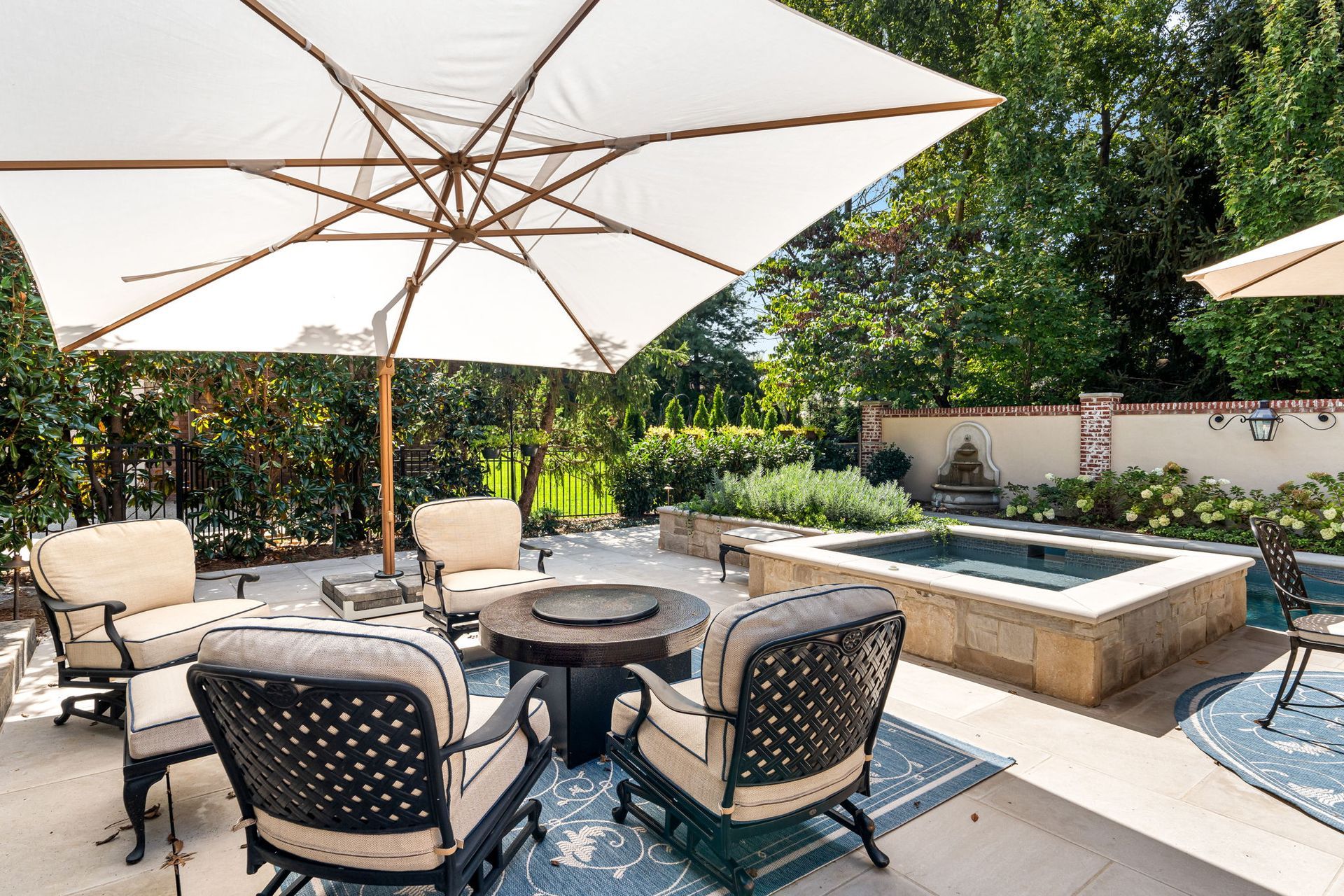 A sunlit patio features a large white umbrella over an outdoor seating area with chairs, a fire pit, and a stone hot tub.