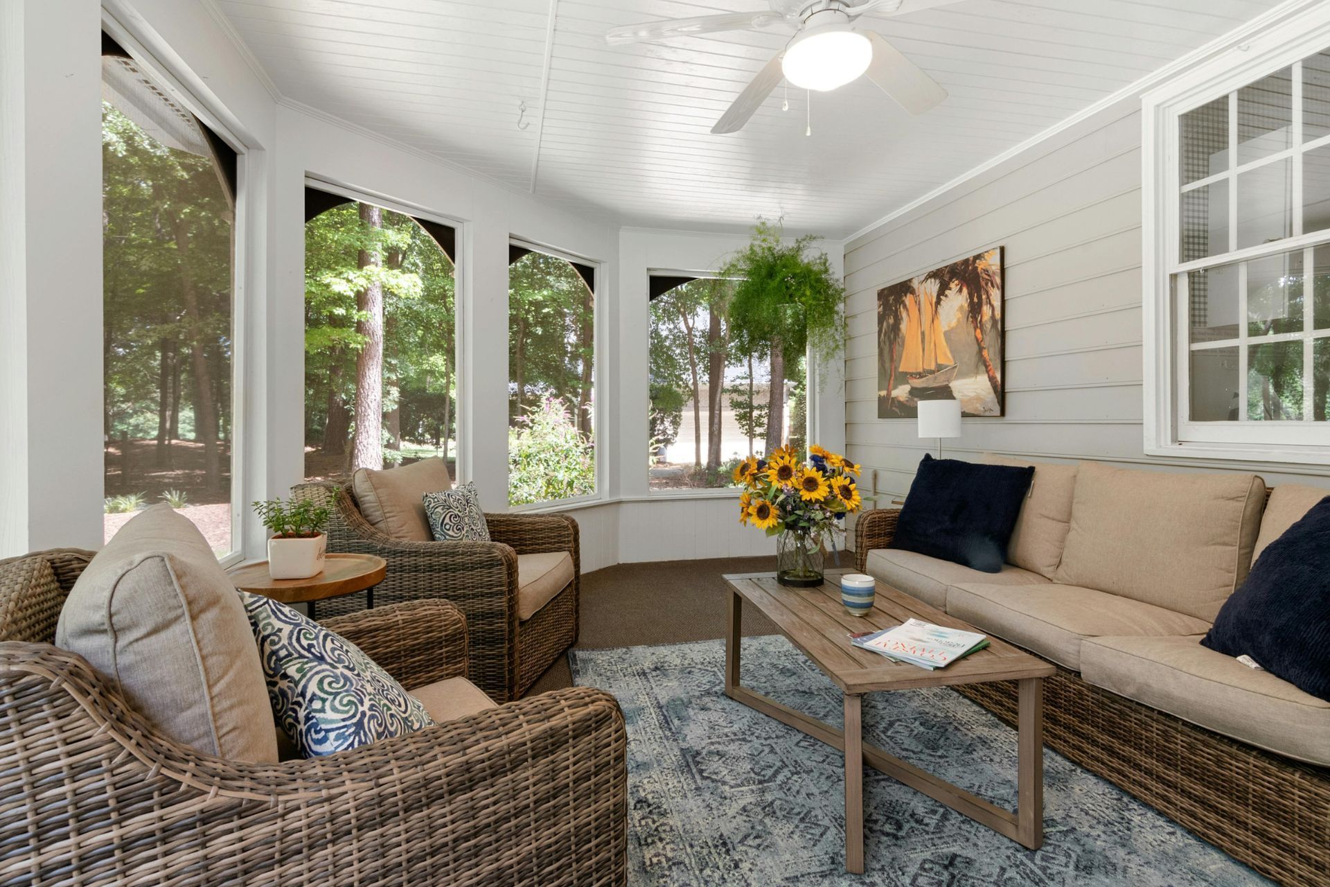 Sunroom with wicker furniture, beige cushions, a coffee table, and large windows overlooking trees.