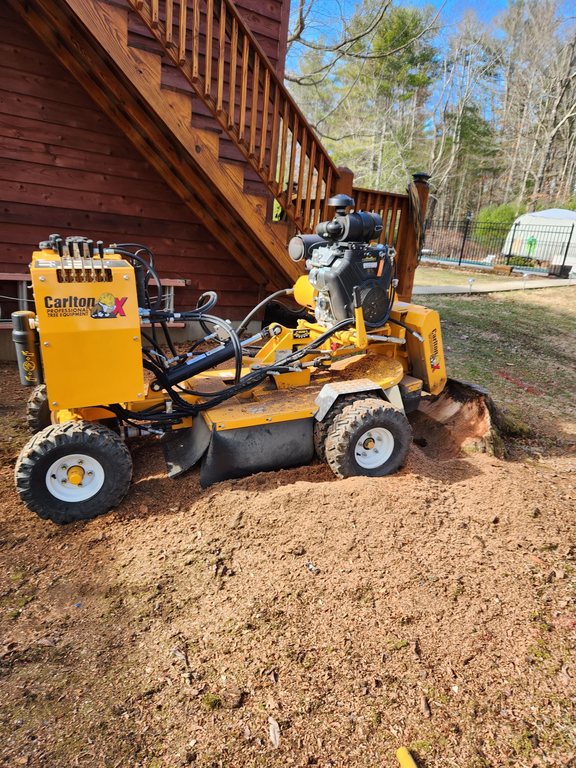 A yellow stump grinder is sitting in the dirt in front of a house.