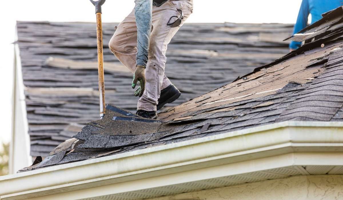 Roofer inspecting a dark roof with a flashlight, wearing a safety harness, yellow hard hat, and kneeling near paperwork.