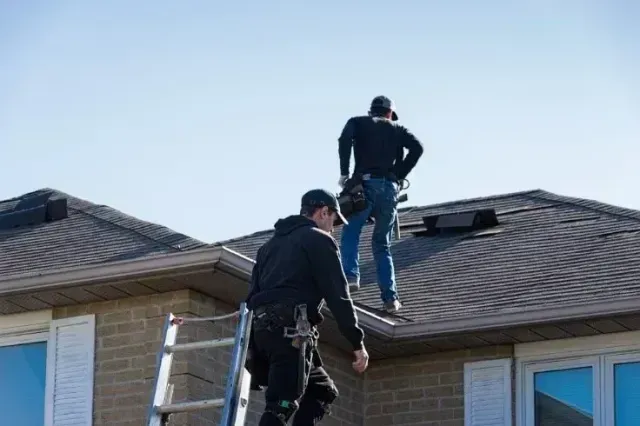 Construction worker on rooftop using a drill. He wears a white vest, hard hat, and holds a power tool.