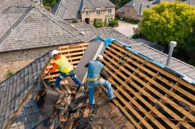 Construction worker on rooftop using a drill. He wears a white vest, hard hat, and holds a power tool.