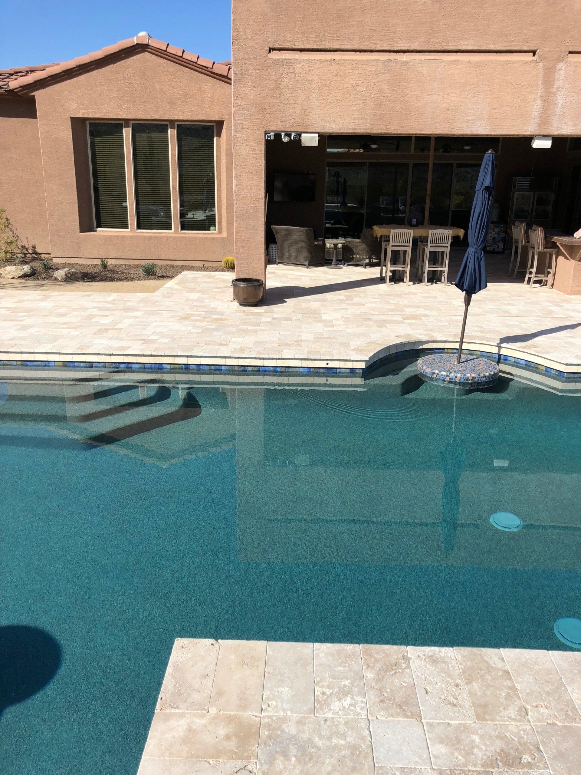 A large swimming pool with stairs and a blue umbrella in front of a house.