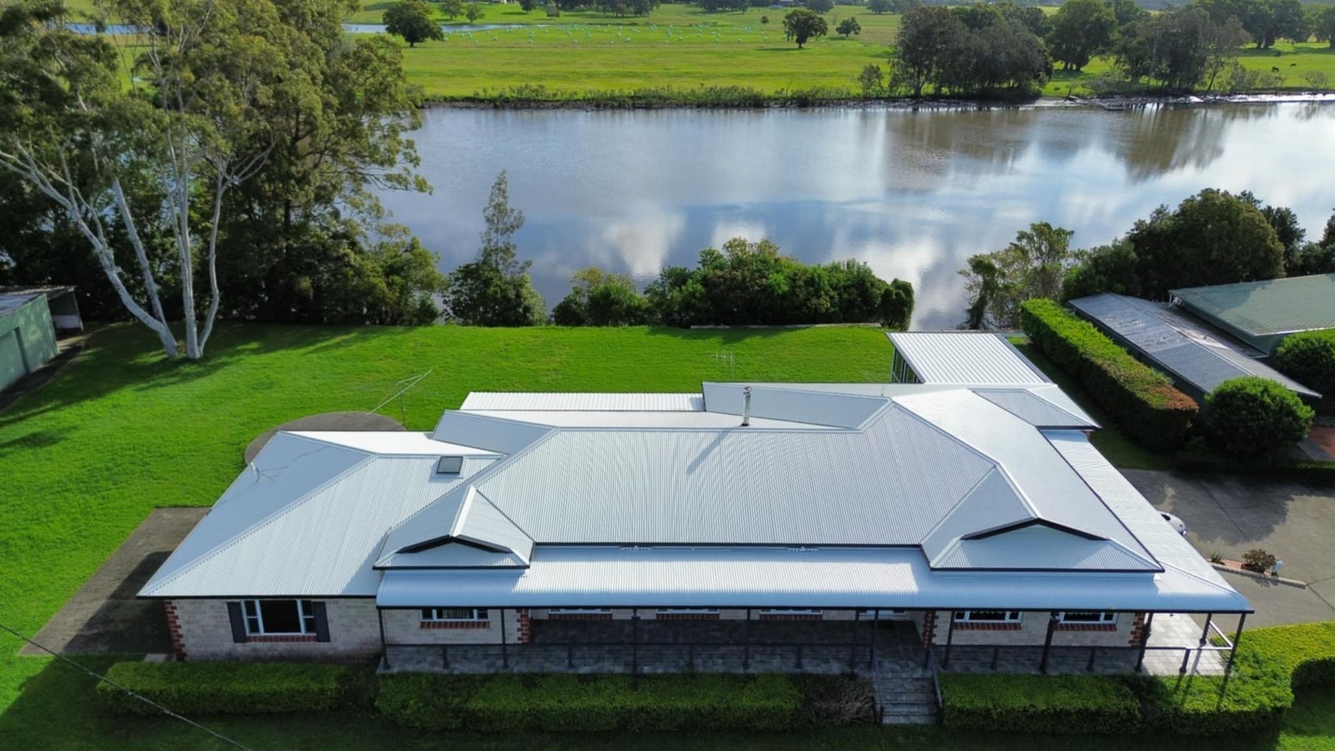 A House With a Large Silver Roof Sits on a Green Lawn Overlooking a River — Tim Leach Metal Roofing in Wauchope, NSW