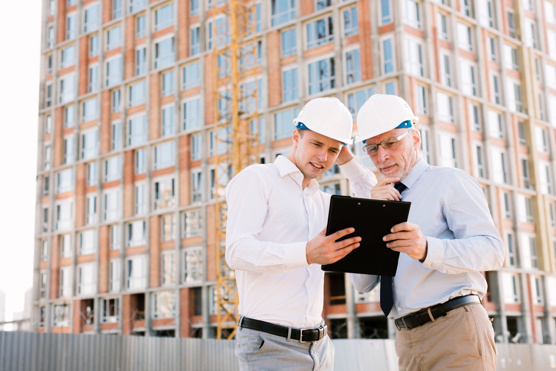 Two men in hard hats are looking at a clipboard in front of a building under construction.