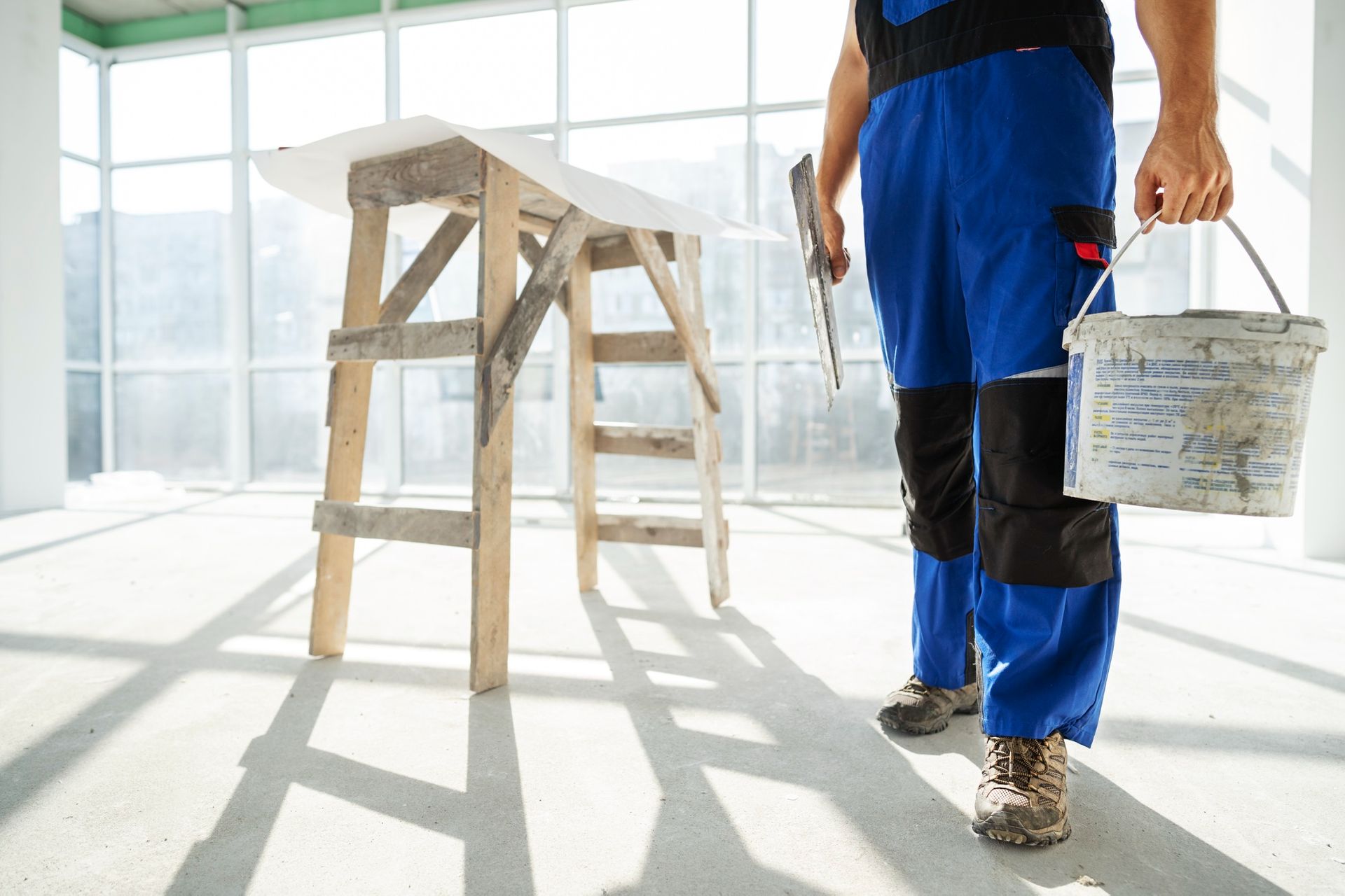 A man in blue overalls is holding a bucket of paint and a spatula.