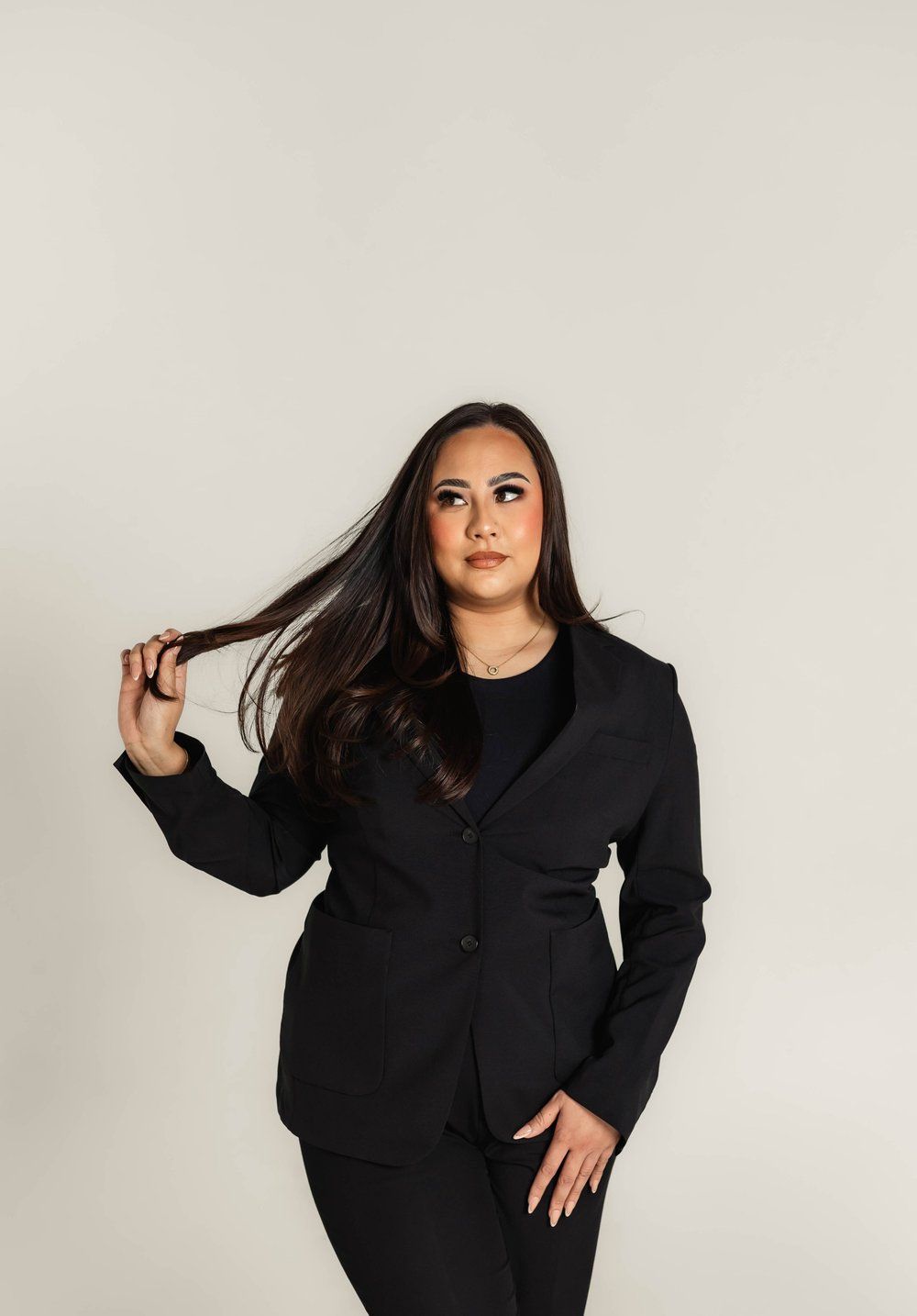 Woman in black suit, looking upwards, holding hair, against a white backdrop.