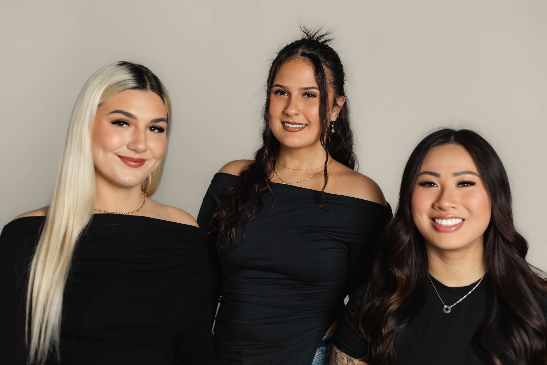 Three women smiling, wearing black off-the-shoulder tops, in front of a neutral background.