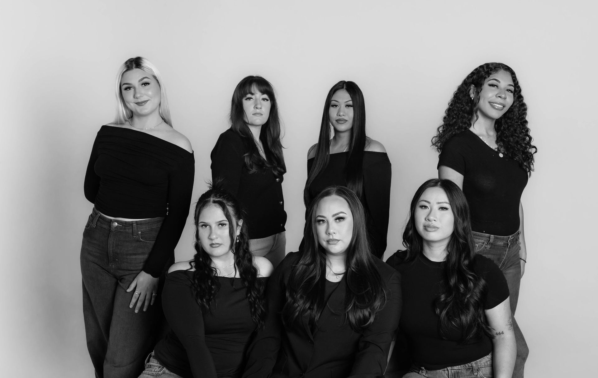 Group of seven people posing together, mostly in black tops and jeans, against a white backdrop.