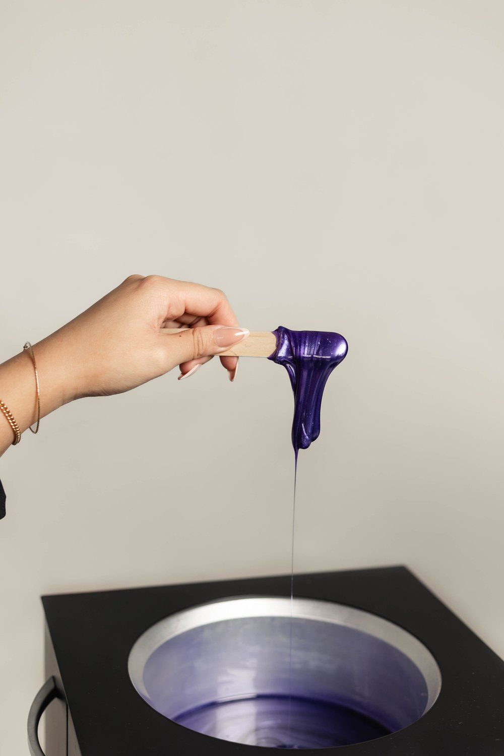 Hand holding a wooden applicator with purple wax, above a wax warmer.