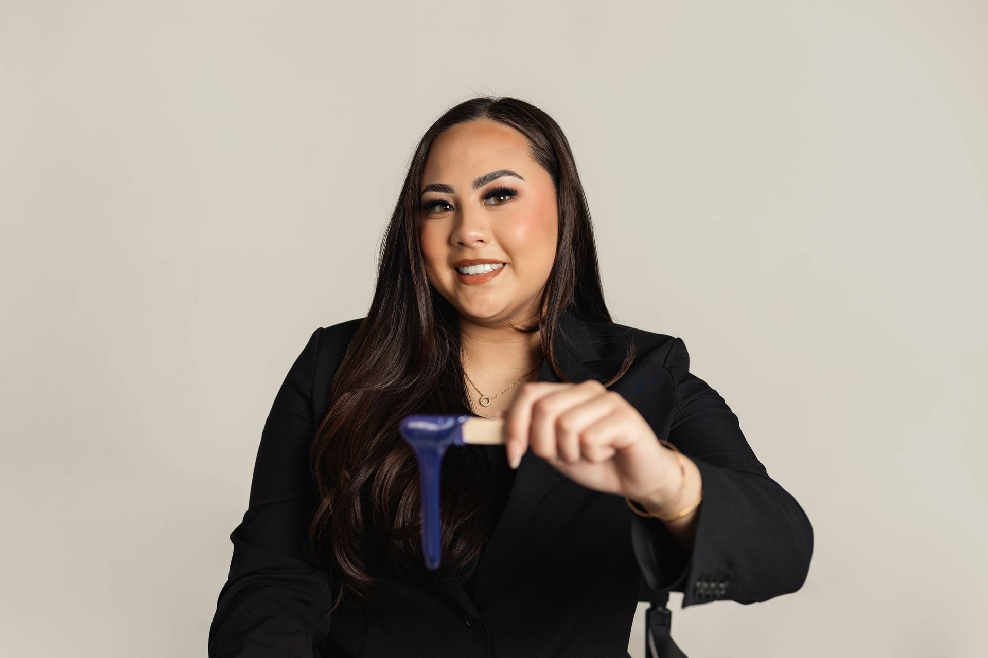 Woman in black blazer smiles, holding a blue and wooden beauty tool against a neutral backdrop.