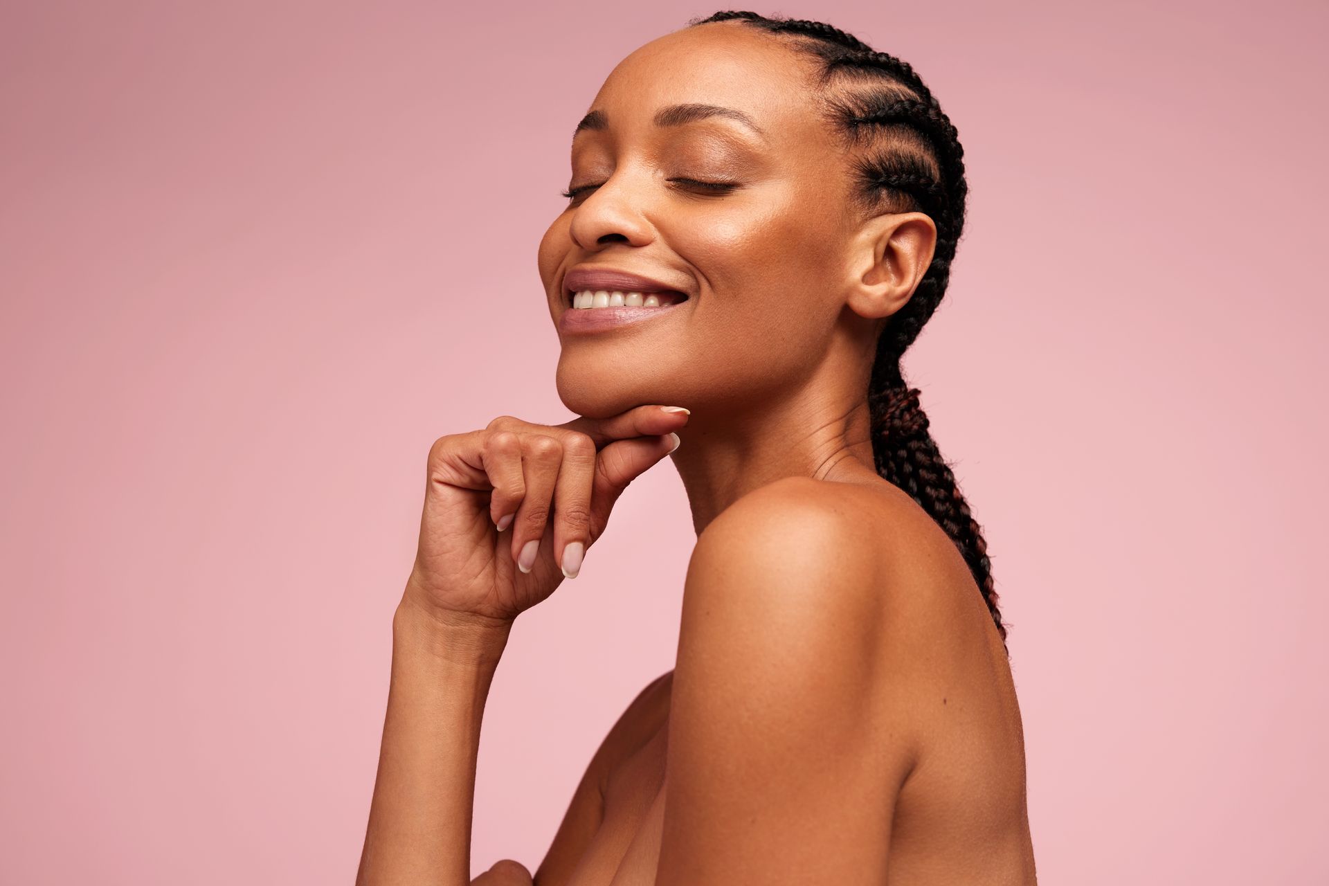 Woman with braided hair smiling, hand on chin, pink background.