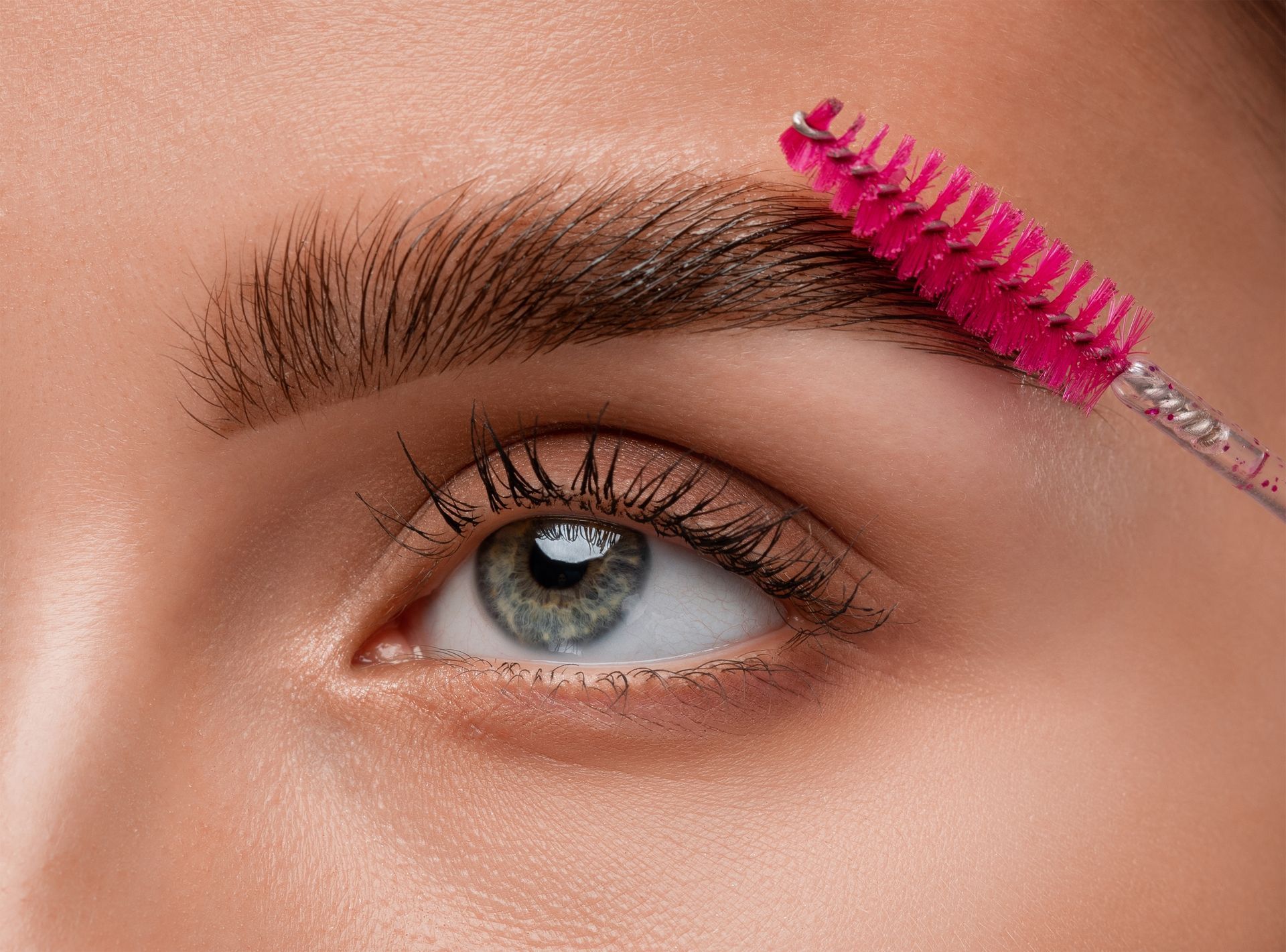 Close-up of eye and eyebrow being brushed with a pink spoolie. The eye is blue-green.