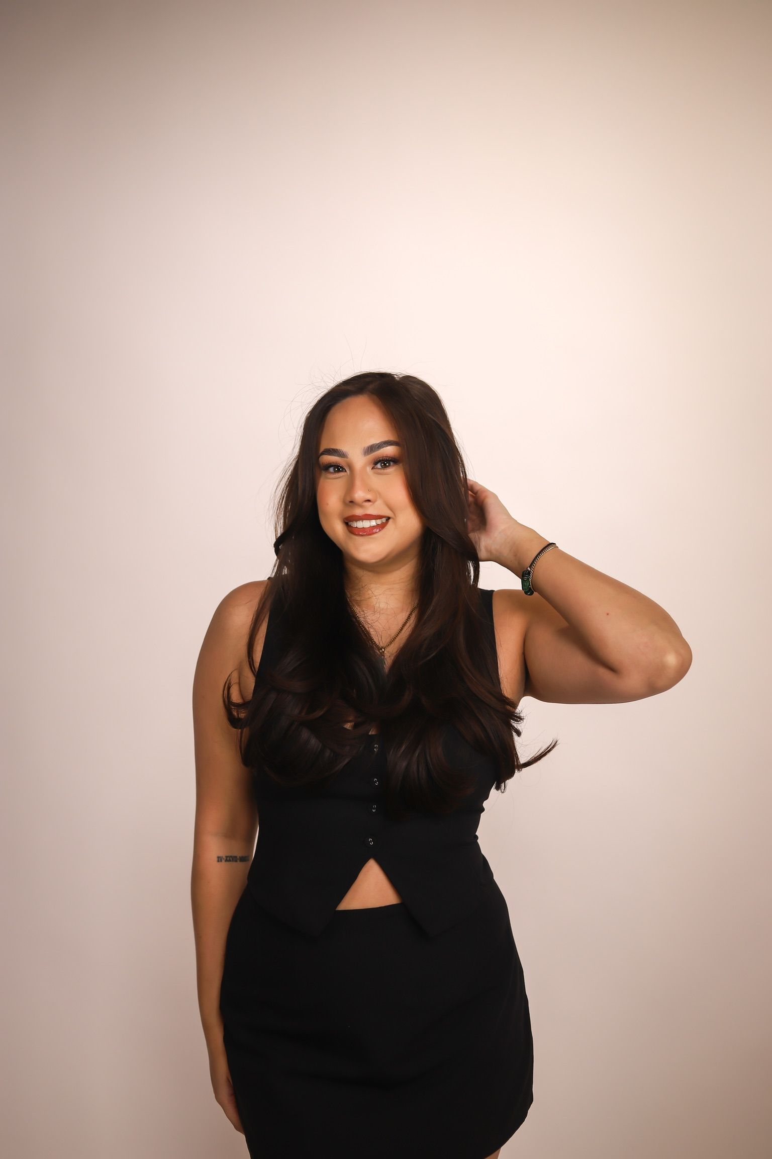 Woman in a black vest and skirt smiles, hand in hair, against a plain background.